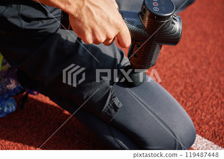 Close-up of male athlete using handheld massage gun on leg muscles for recovery after training session on stadium track. Concept of muscle relaxation and sports recovery 119487448