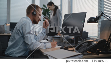 In modern office: Financial trader in headset works on computer, communicates with client 119489200