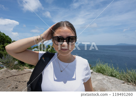 A close-up portrait of a smiling teenage girl wearing sunglasses. Ocean view in the background. 119491260