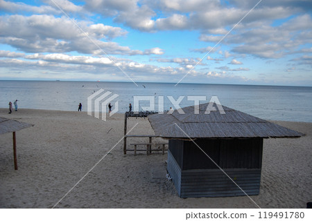 Landscape of Odessa beach. Odessans stroll along the autumn beach and feed seagulls. A cafe with a thatched roof and an improvised bar counter is empty. Landscape of Odessa beach. Odessans stroll along the autumn beach and feed seagulls. A cafe with a thatched roof and an improvised bar counter is empty. 119491780