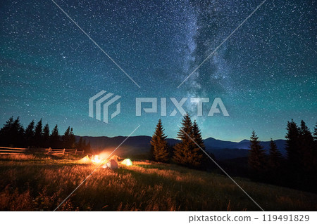 Silhouettes of tourists resting at campsite around campfire with glowing tents nearby under starry sky. Milky Way stretches above, casting serene and magical light over tranquil mountain landscape. 119491829