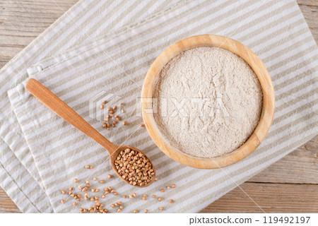 Buckwheat flour in a wooden bowl with wooden scoop on beige a cotton napkin, top view 119492197