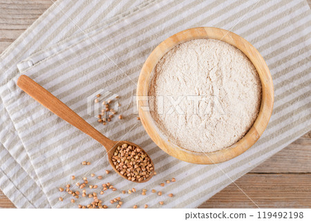 Buckwheat flour in a wooden bowl with wooden scoop on beige a cotton napkin, top view 119492198