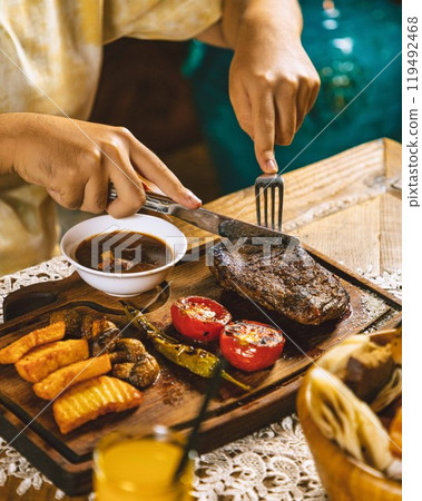Woman cutting grilled beef steak served on wooden board with vegetables Woman cutting grilled beef steak served on wooden board with vegetables 119492468