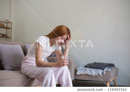 Woman Sitting on Sofa Holding Head in Pain, Suffering from Headache or Stress, Indoor Home Setting 119493562