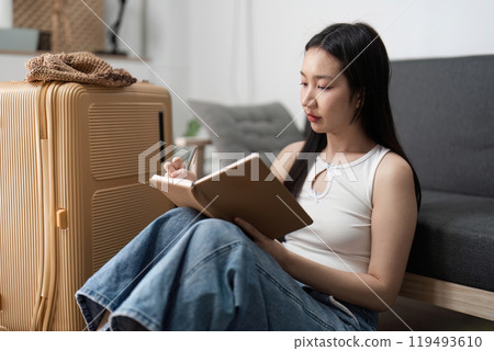 Young Woman Preparing for Travel by Packing Suitcase and Making Notes at Home 119493610