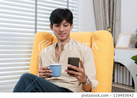 Young Man Relaxing in Modern Room, Sitting on Yellow Chair, Using Smartphone and Drinking Coffee 119493642