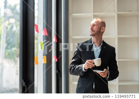 Young Businessman with Determination Looking Out Window Holding Coffee Cup in Modern Office Setting Young Businessman with Determination Looking Out Window Holding Coffee Cup in Modern Office Setting 119493771