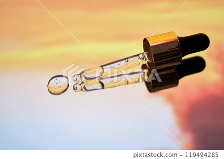A pipette with facial serum against a sunset background in bright orange tones. 119494285