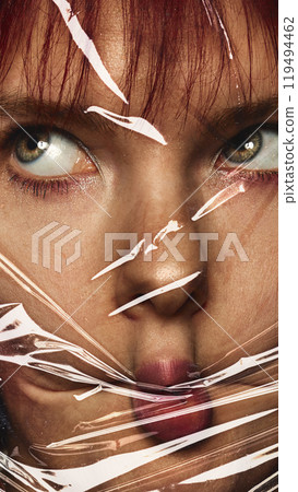 Close-up of female face wrapped in plastic cover. Woman making funny, grimacing face. Conceptual photography. 119494462