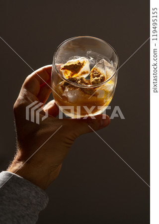 A glass filled with ice cubes and powdered Amanita muscaria mushroom on its surface, held in the hand, illuminated by natural sunlight. Preparing a homemade fly agaric elixir 119495155
