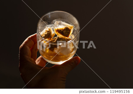 A glass filled with ice cubes and powdered Amanita muscaria mushroom on its surface, held in the hand, illuminated by natural sunlight. Preparing a homemade fly agaric elixir 119495156