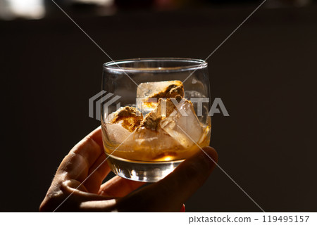 A glass filled with ice cubes and powdered Amanita muscaria mushroom on its surface, held in the hand, illuminated by natural sunlight. Preparing a homemade fly agaric elixir 119495157