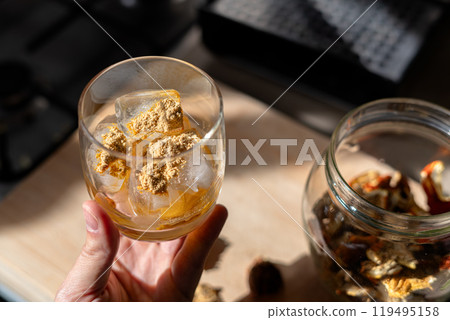 A glass filled with ice cubes and powdered Amanita muscaria mushroom on its surface, held in the hand, illuminated by natural sunlight. Preparing a homemade fly agaric elixir A glass filled with ice cubes and powdered Amanita muscaria mushroom on its surface, held in the hand, illuminated by natural sunlight. Preparing a homemade fly agaric elixir 119495158