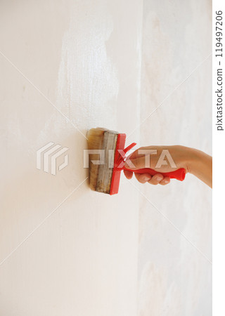 Person is holding brush with glue on background of light wall. apply thick wallpaper glue.  119497206