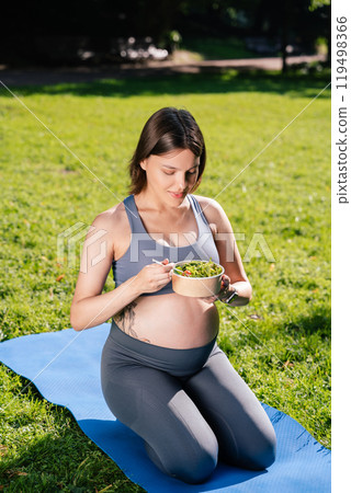 Pregnant woman holding plate with salad eating after yoga exercising sitting on green grass 119498366