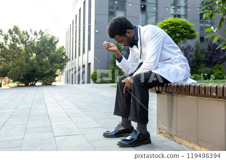 Exhausted doctor man in medical gown with stethoscope sitting outside near hospital 119498394