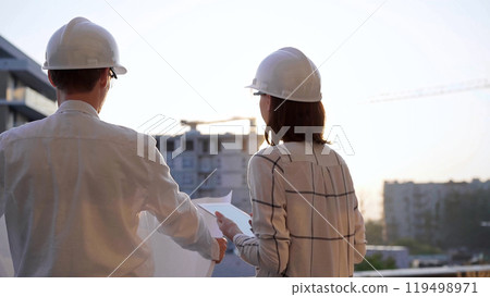 Two architects, wearing hard hat and safety glasses, are examining blueprints at a construction site during sunrise or sunset, viewed from the back. Architecture and engineering 119498971