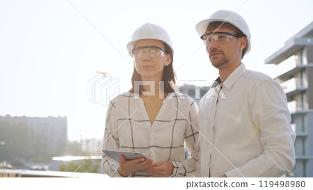 Two architects colleagues, wearing safety helmets and glasses, are standing near each other and discussing a construction site progress during sunrise or sunset. Architecture and engineering Two architects colleagues, wearing safety helmets and glasses, are standing near each other and discussing a construction site progress during sunrise or sunset. Architecture and engineering 119498980