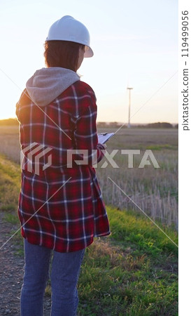 Woman engineer wearing a white protective helmet is taking notes with a clipboard in a field with wind turbines, as the sun sets. Clean energy and engineering concept 119499056