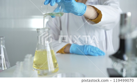 A scientist, wearing a lab coat and blue protective gloves, is pouring a yellow liquid from one beaker to another in laboratory, close up. Medicine and science concepts 119499070