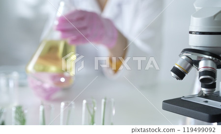 Microscope is near a scientist, wearing a lab coat and pink protective gloves, and holding beaker with a yellow oily liquid in laboratory setting, close up. Medicine and science 119499092