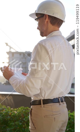 Man a construction engineer wearing white shirt and hard hat is making notes on a clipboard while inspecting a building site at sunset, back vertical view . Architecture concept Man a construction engineer wearing white shirt and hard hat is making notes on a clipboard while inspecting a building site at sunset, back vertical view . Architecture concept 119499119