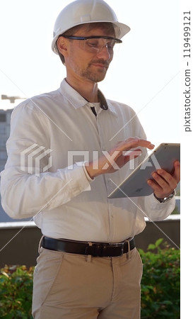 Male construction engineer wearing white shirt and helmet is using digital tablet while inspecting a building site at sunrise, front and vertical view . Architecture concept 119499121