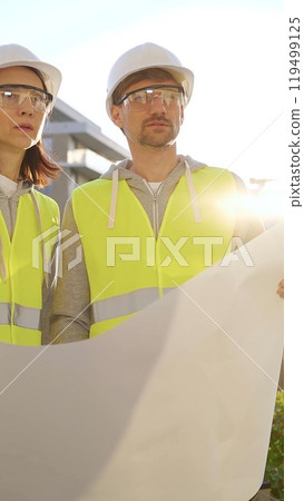 Two architects or engineers wearing safety hard hats and vests holding blueprint and discussing something on construction site at sunrise, front vertical view. Architecture engineering and teamwork 119499125