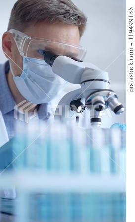 Man scientist with protective glasses, mask and gloves is using microscope in laboratory, vertical portrait of researcher. Microbiology and science Man scientist with protective glasses, mask and gloves is using microscope in laboratory, vertical portrait of researcher. Microbiology and science 119499136
