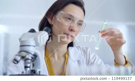 Scientist researcher with protective glasses examining lab tube with a green fluid. Microbiology and science concept Scientist researcher with protective glasses examining lab tube with a green fluid. Microbiology and science concept 119499137