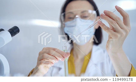 Woman with safety glasses and blue mask is working with lab tubes using a pipette in a modern laboratory, close-up of researcher. Science and medicine Woman with safety glasses and blue mask is working with lab tubes using a pipette in a modern laboratory, close-up of researcher. Science and medicine 119499178