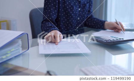 A female accountant with blue dotted blousy is using a laptop computer and calculator to calculate taxes at a glass desk in the office A female accountant with blue dotted blousy is using a laptop computer and calculator to calculate taxes at a glass desk in the office 119499190