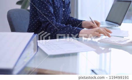 A female accountant with blue dotted blousy is using a laptop computer and calculator to calculate taxes at a glass desk in the office. Right side view A female accountant with blue dotted blousy is using a laptop computer and calculator to calculate taxes at a glass desk in the office. Right side view 119499191