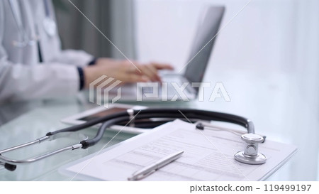 Clipboard with medical records and stethoscope are lying on the glass table while doctor woman is using tablet computer on the glass table. Medicine concept 119499197