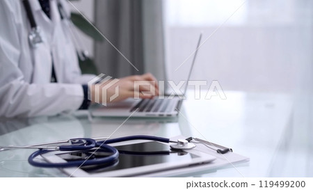 Tablet computer and blue stethoscope are on the glass table while doctor woman is using laptop computer on the glass table. Medicine concept Tablet computer and blue stethoscope are on the glass table while doctor woman is using laptop computer on the glass table. Medicine concept 119499200