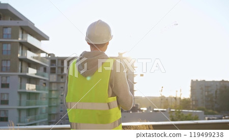 Male engineer with white hard hat and safety vest is using a tablet computer while inspecting a construction site at sunset 119499238