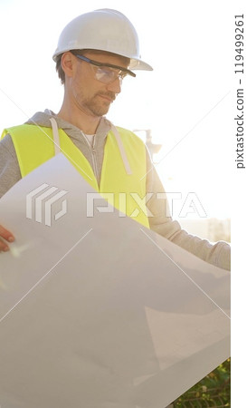Man an architect or engineer wearing safety hard hat and vest holding blueprint and looking to construction site at sunrise, front vertical view. Architecture concept Man an architect or engineer wearing safety hard hat and vest holding blueprint and looking to construction site at sunrise, front vertical view. Architecture concept 119499261