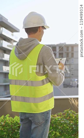 Man engineer with white helmet and safety vest is taking notes on a clipboard while inspecting a construction site at sunrise, back vertical view Man engineer with white helmet and safety vest is taking notes on a clipboard while inspecting a construction site at sunrise, back vertical view 119499314