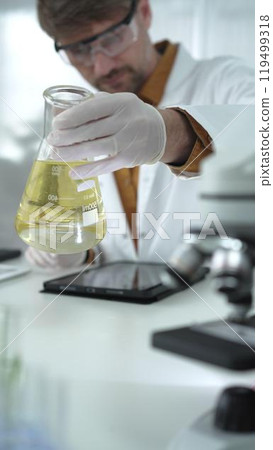 Man scientist, wearing white gloves and protective glasses, is holding a yellow chemical solution inside Erlenmeyer flask near microscope in a laboratory, vertical portrait view. Science and medicine Man scientist, wearing white gloves and protective glasses, is holding a yellow chemical solution inside Erlenmeyer flask near microscope in a laboratory, vertical portrait view. Science and medicine 119499318