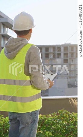 Man engineer with white helmet and safety vest is taking notes on a clipboard while inspecting a construction site at sunrise, back vertical view 119499321