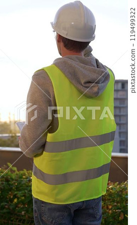 Male engineer with white helmet and safety vest is taking notes on a clipboard while inspecting a construction site at sunrise, vertical back view. Architecture and engineering Male engineer with white helmet and safety vest is taking notes on a clipboard while inspecting a construction site at sunrise, vertical back view. Architecture and engineering 119499322