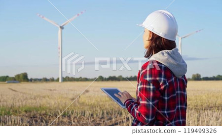 Female engineer wearing a white protective helmet is taking notes with a tablet computer on a field with wind turbines, as the sun sets. Clean energy and engineering 119499326