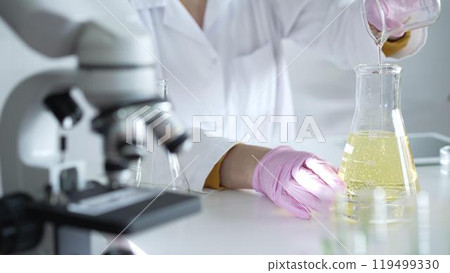 A scientist, wearing a lab coat and pink protective gloves, is pouring a yellow oily liquid from one beaker to another near microscope in laboratory, close up. Medicine and science A scientist, wearing a lab coat and pink protective gloves, is pouring a yellow oily liquid from one beaker to another near microscope in laboratory, close up. Medicine and science 119499330