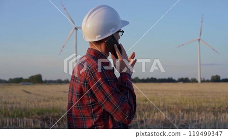 Man engineer, wearing a white protective helmet is talking by smartphone in a field with wind turbines, as the sun sets. Clean energy and engineering concept 119499347