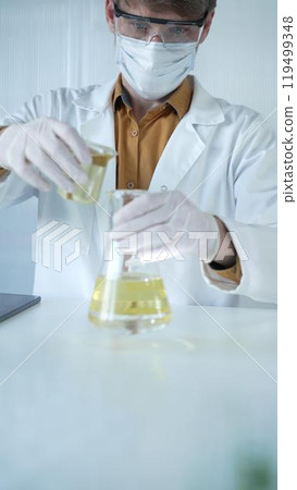Man scientist wearing a lab coat, white gloves, mask, and protective glasses, is pouring a yellow oily liquid from one beaker to another in a laboratory setting, close up. Science and medicine Man scientist wearing a lab coat, white gloves, mask, and protective glasses, is pouring a yellow oily liquid from one beaker to another in a laboratory setting, close up. Science and medicine 119499348