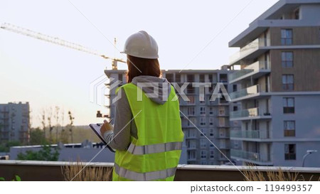 Construction female engineer taking notes and writing on clipboard while inspecting a building site at sunset. Industry development concept 119499357