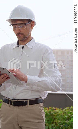 Male architect, wearing a white shirt and helmet, is using a digital tablet while inspecting a building site in foggy weather. Architecture and engineering concept 119499358