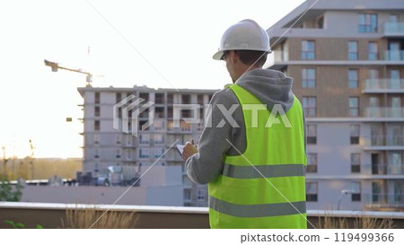Man engineer with a white protective helmet and safety vest is writing on a clipboard while inspecting a construction site in early morning at sunrise. Architect concept 119499366