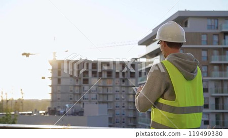 Man constructive engineer with white hard hat and safety vest is using a tablet computer while inspecting a construction site at sunset 119499380